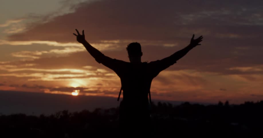 A silhouetted hiker with a backpack slowly raises his arms wide to embrace a beautiful golden sunset over the ocean