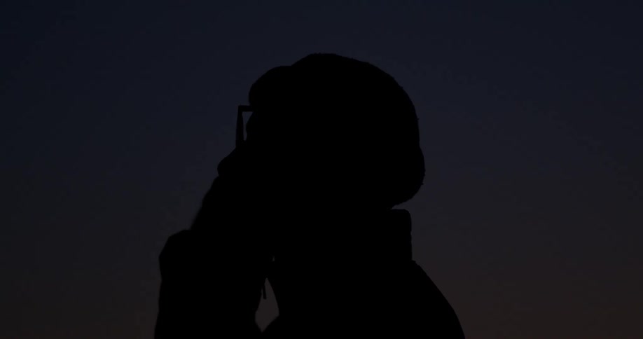 The silhouette of a man wearing a beanie and glasses talks on the phone at dusk