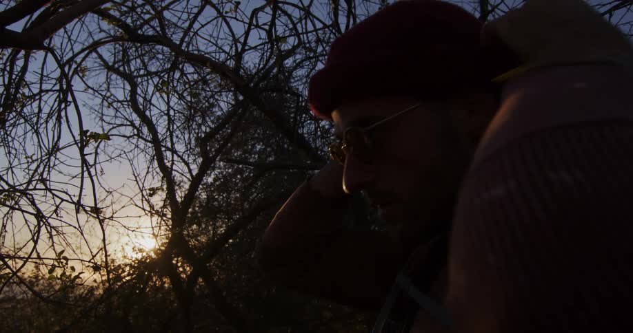 A young man with a pencil behind his ear puts on his red beanie while hiking at sunset, silhouetted against the trees