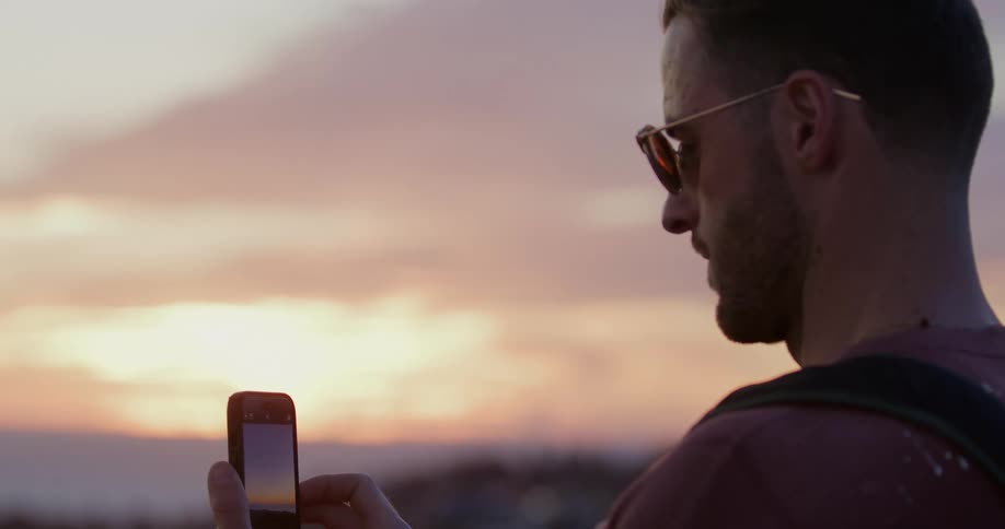 A man takes a picture of a beautiful sunset with his smartphone, then looks up to admire the serene view