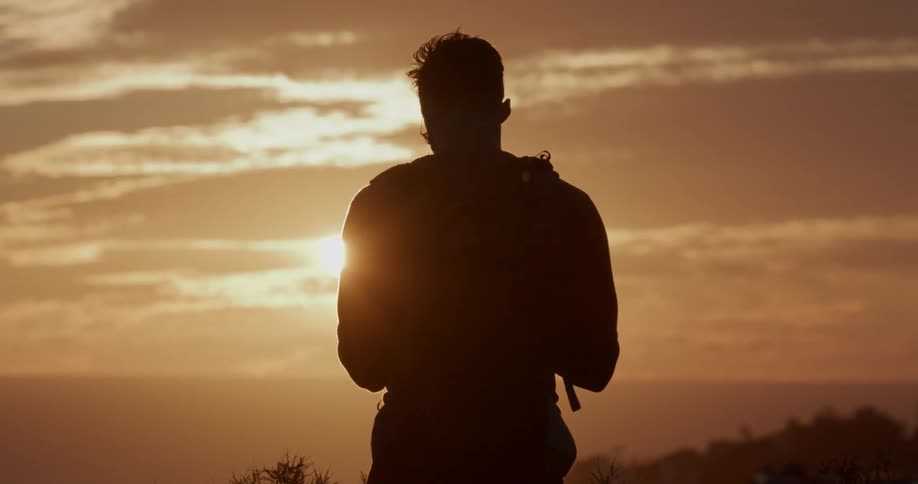 A silhouetted hiker with a backpack watches the golden sunset over the ocean from a scenic viewpoint