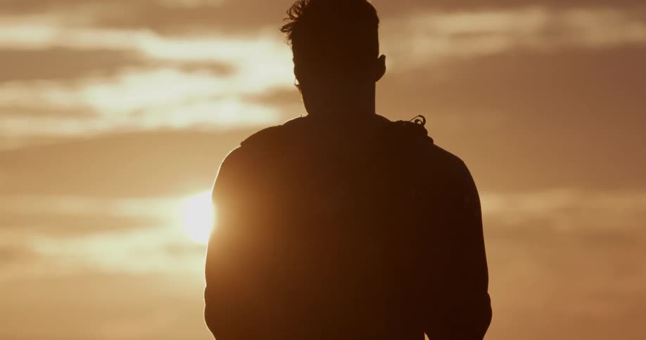 Back view of a silhouetted man standing still while looking at the bright sun during golden hour