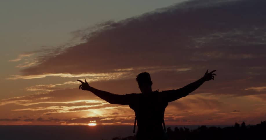 A man's silhouette stands with arms raised in slow motion, embracing the magnificent sunset over the tranquil ocean
