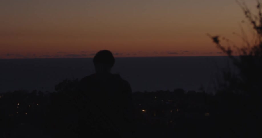 A silhouetted hiker walks toward a scenic overlook to watch the sunset over a coastal city and the dark ocean