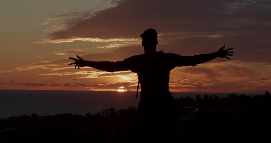 A silhouetted person with a backpack raises their arms in slow motion to embrace a glorious sunset over the ocean
