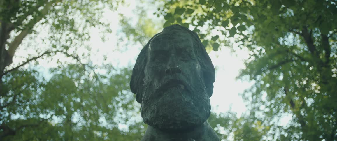 A low-angle shot of the weathered bust of Karl Marx, surrounded by a canopy of green trees in a park