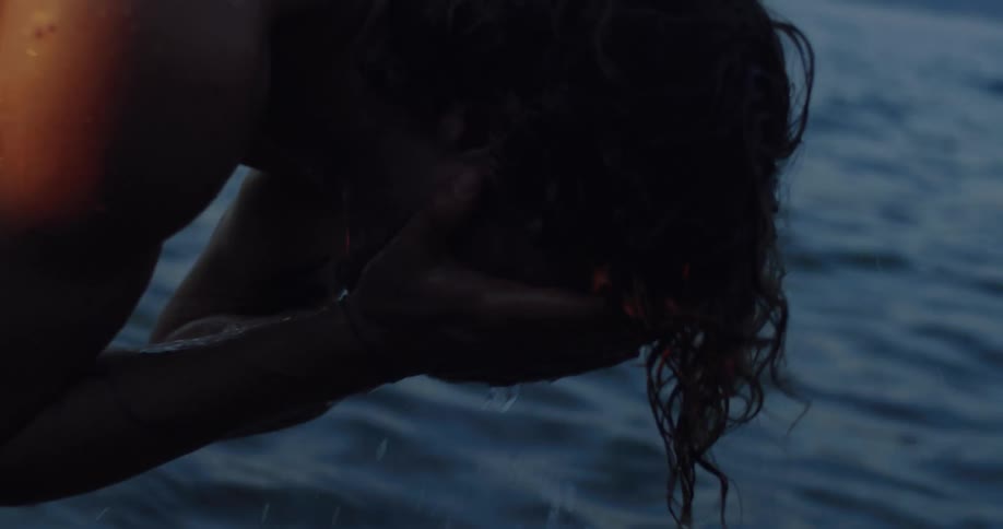 A shirtless man with long curly hair splashes his face with cool water from the sea at dusk in slow motion