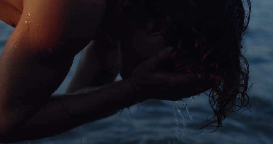 Slow motion close-up of a person with curly hair washing their face in the cool ocean water at dusk