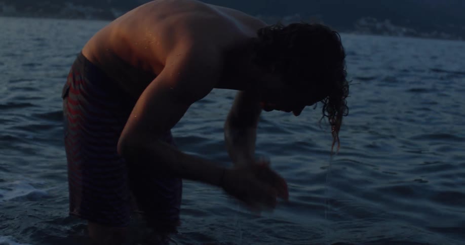 A young man with long hair stands in the ocean at dusk, repeatedly splashing his face with water in slow motion