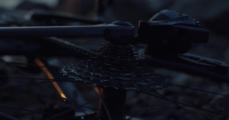 A close-up shot captures the rear cassette and chain of a bicycle spinning slowly while lying on the ground at dusk