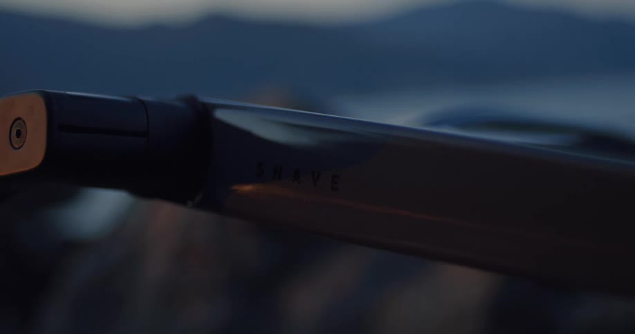A close-up of a sleek, modern razor handle with the word 'SHAVE' inscribed on it against a blurred natural background at dusk