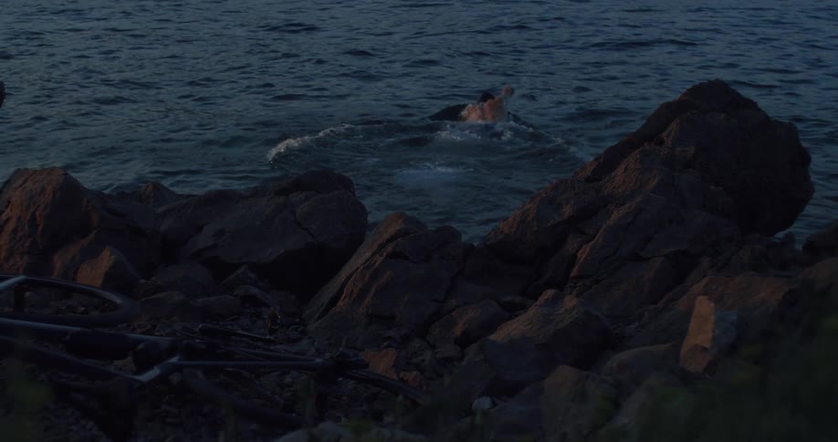 A shirtless man dives backward in slow motion into the water from a rocky shore at twilight and begins to swim