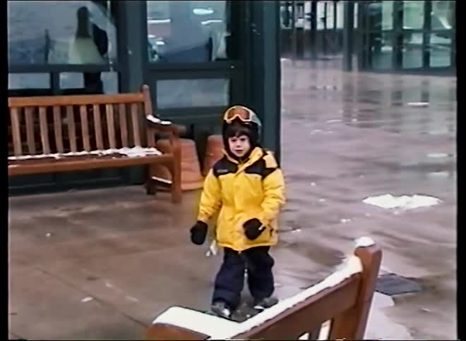 A vintage home video shows a young boy dressed in a bright yellow ski jacket and helmet at a snowy resort