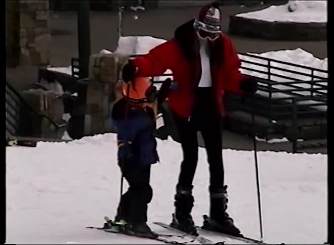 A mother helps her young child stand up on skis for the first time in this vintage footage from a ski resort