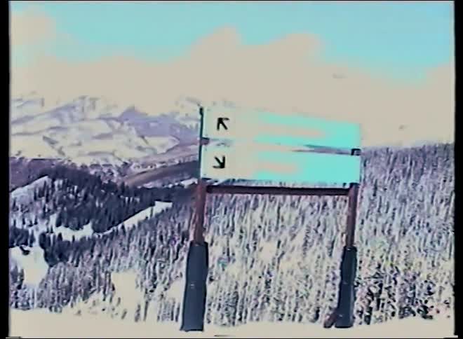 A vintage point-of-view shot pans across a vast, snowy mountain range at a ski resort, revealing a trail sign