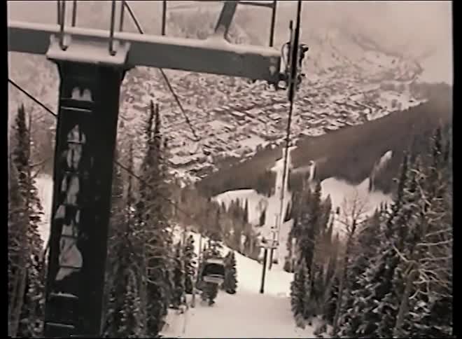 Vintage point-of-view footage from a ski gondola descending a snow-covered mountain, overlooking a small resort town in the valley below