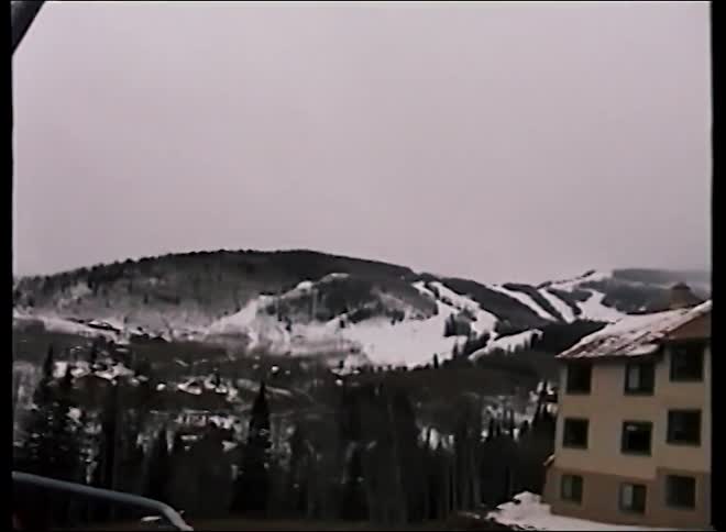 Vintage VHS footage shows a person on a ski lift looking out at the snowy mountain slopes and forest below on an overcast day