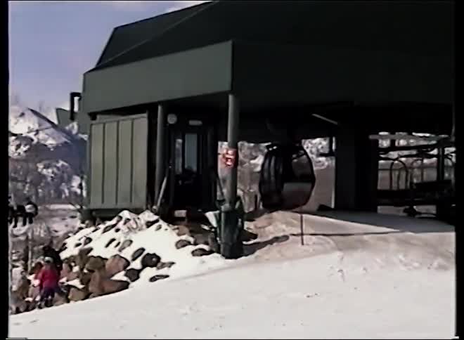 A vintage VHS recording pans across a bustling ski resort with skiers, chairlifts, and a gondola station against a backdrop of majestic snowy mountains