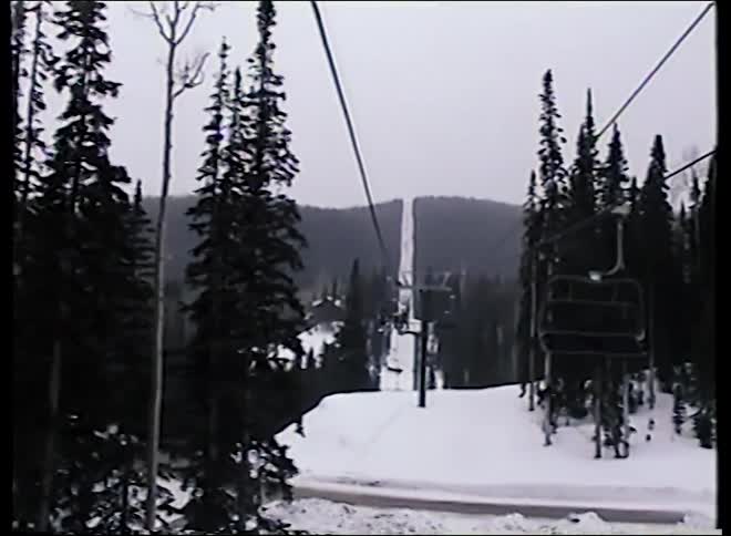 A point-of-view shot from a ski lift ascending a snowy mountain covered in evergreen trees on an overcast day