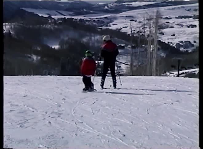 A young child learns to ski with an adult instructor on a snowy mountain slope with a vast valley below