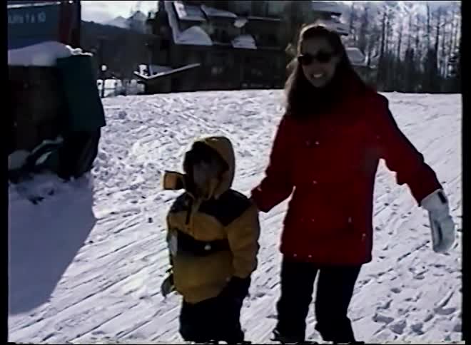 A vintage home video shows a smiling mother teaching her young child how to ski on a sunny day at a mountain resort