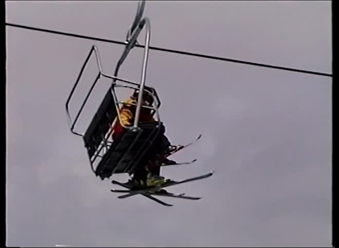Vintage footage shows three skiers riding a chairlift up a snowy mountain resort on an overcast day