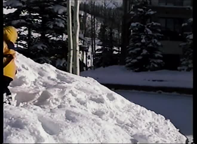 A vintage home movie shows a toddler playing in the snow, then pans to a woman in a red jacket smiling and posing at a winter resort