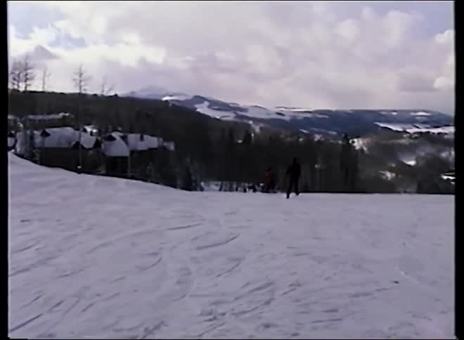 Vintage home video footage shows skiers on a wide, snowy slope with a stunning mountain valley in the background