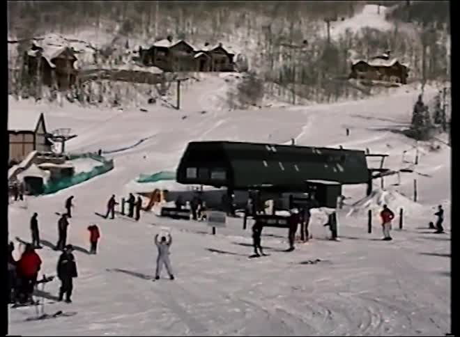 Wide shot of a bustling ski resort pans to a woman in a purple snowsuit joyfully waving her gloves in this vintage footage