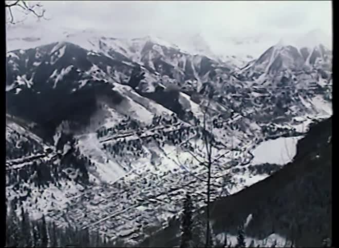 A vintage, high-angle pan across a vast, snow-covered mountain range reveals a small town and frozen lake in the valley