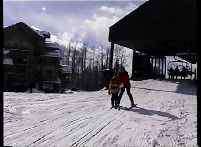 Vintage home movie footage captures a mother teaching her young child how to ski on a snowy mountain slope