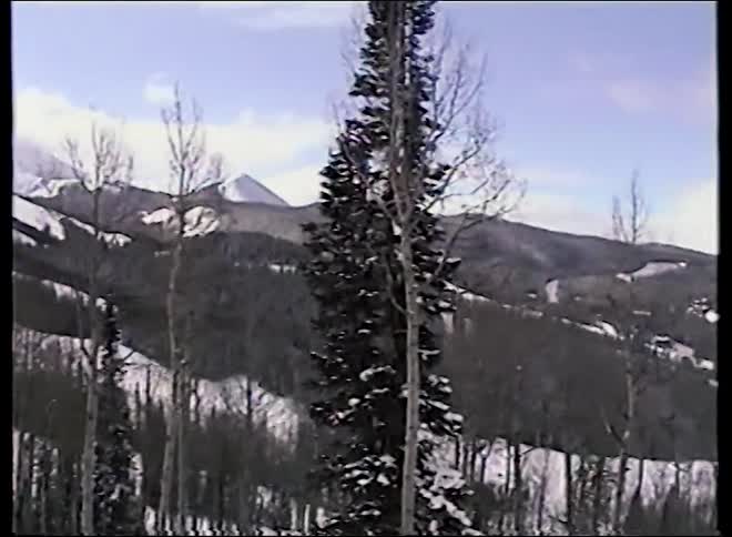 Vintage footage slowly pans across a tranquil, snowy mountain range revealing ski slopes behind a winter forest