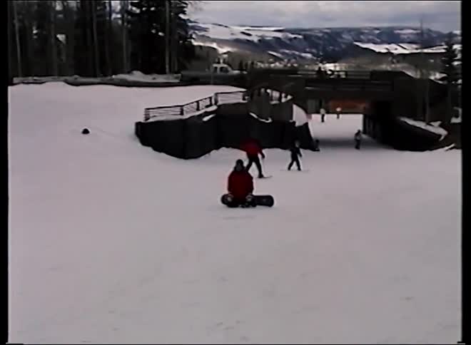 A vintage home video shows a beginner snowboarder in a red jacket falling while learning to ride down a snowy mountain slope