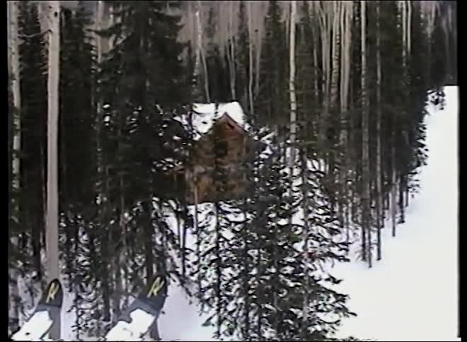 A point-of-view shot from a moving ski lift reveals a beautiful log cabin nestled in a snowy, wooded mountain landscape