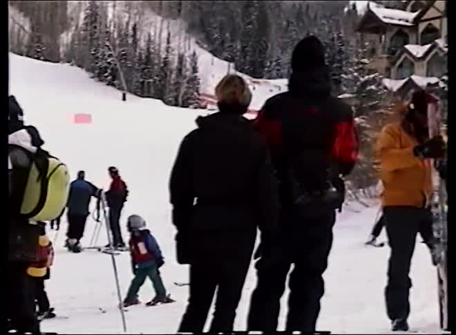 Archival footage shows a group of skiers and snowboarders gathered on a snowy mountain slope at a busy ski resort