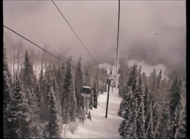 Vintage footage from a gondola shows a point-of-view ride over a snowy mountain forest on a foggy day