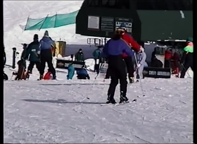 Vintage footage follows a skier down a gentle slope towards a bustling ski lift station at a mountain resort