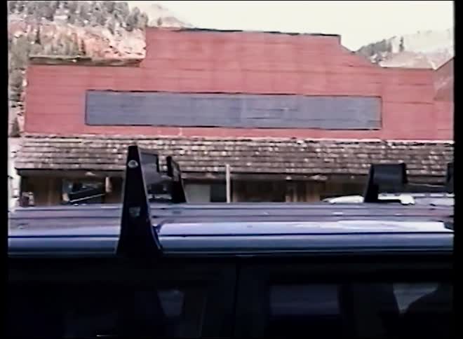 Vintage footage pans across a quaint mountain town, showing a street with a clock tower set against a snowy peak and colorful Victorian homes
