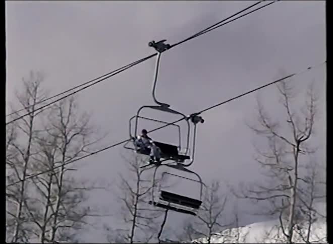 A lone skier rides a chairlift up a snowy mountain, passing bare trees on an overcast day