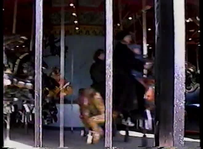 A close-up shot of a painted horse decoration on a fence pans up to reveal a classic carousel spinning with riders