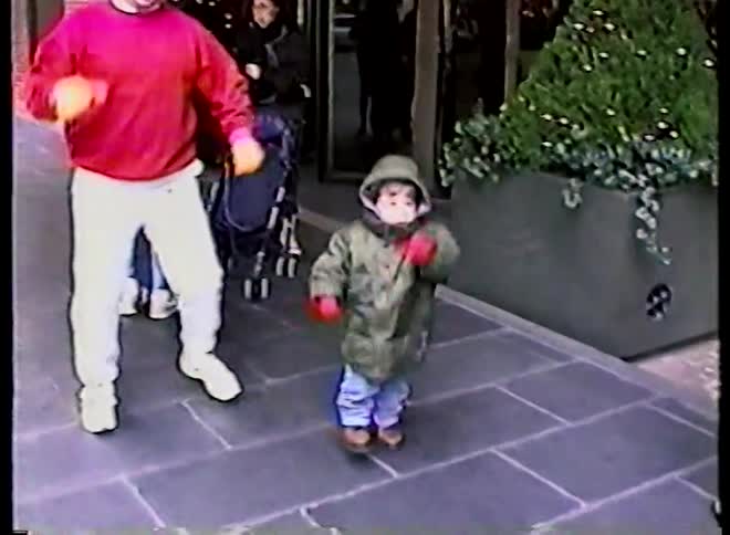 A joyful toddler in a puffy winter coat dances on a city sidewalk while their father playfully dances alongside