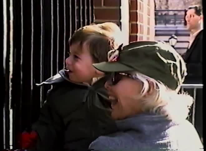 A smiling woman holds a young boy as they both look with wonder and excitement at something off-screen, possibly at a zoo or park