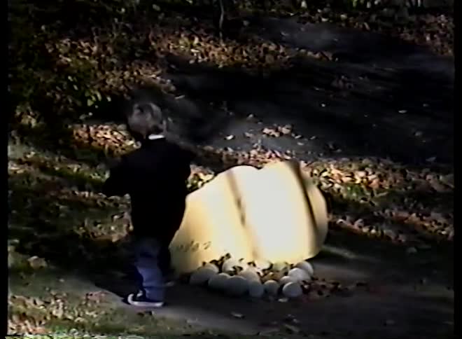 Vintage home movie footage captures a little boy discovering and playing with a large, cracked dinosaur egg sculpture in a park