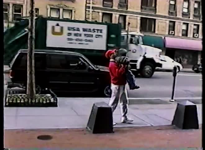 Vintage home movie footage shows a father lifting his young child onto his shoulders on a busy city street