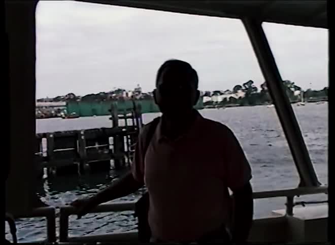 A silhouetted man stands on a water taxi as it departs from a harbor on a cloudy day, captured on vintage VHS
