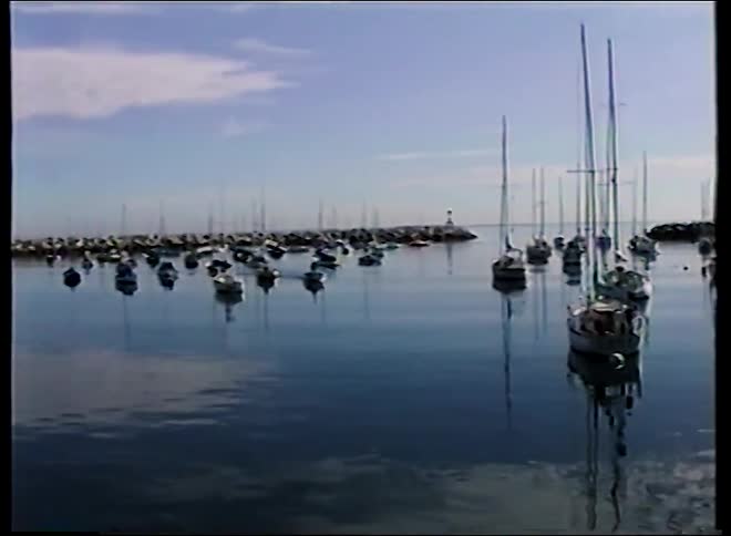 A slow pan reveals numerous sailboats and yachts moored in a calm, reflective harbor on a sunny day