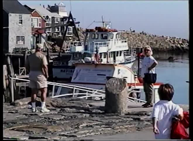 A family stands on a rustic pier in a quaint fishing harbor on a sunny day, captured on vintage home video