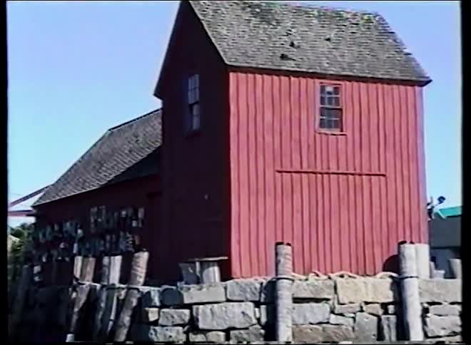 A seagull perches on the chimney of a classic red fishing shack before the camera pans to reveal a quaint New England harbor