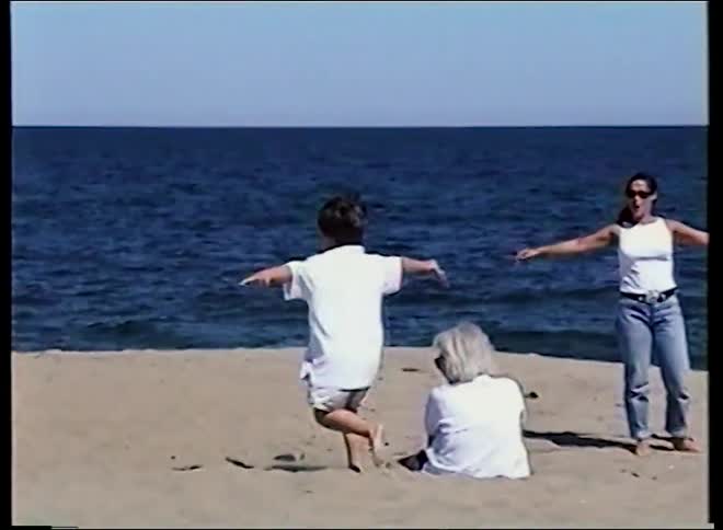 A young boy runs in slow motion with his arms outstretched on a sandy beach while his mother and grandmother watch