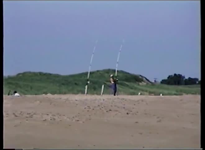 Vintage home video of a person surf fishing on a sandy beach with large dunes in the background